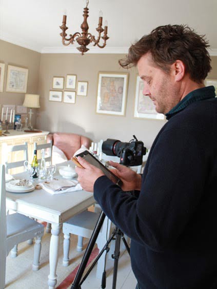 Man holding a camera and checking settings while standing next to a tripod in an interior space.