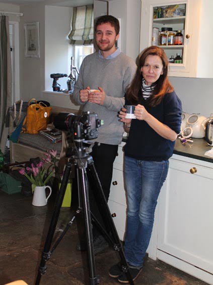 Two individuals standing beside a camera on a tripod, one holding a mug and the other holding a cup.
