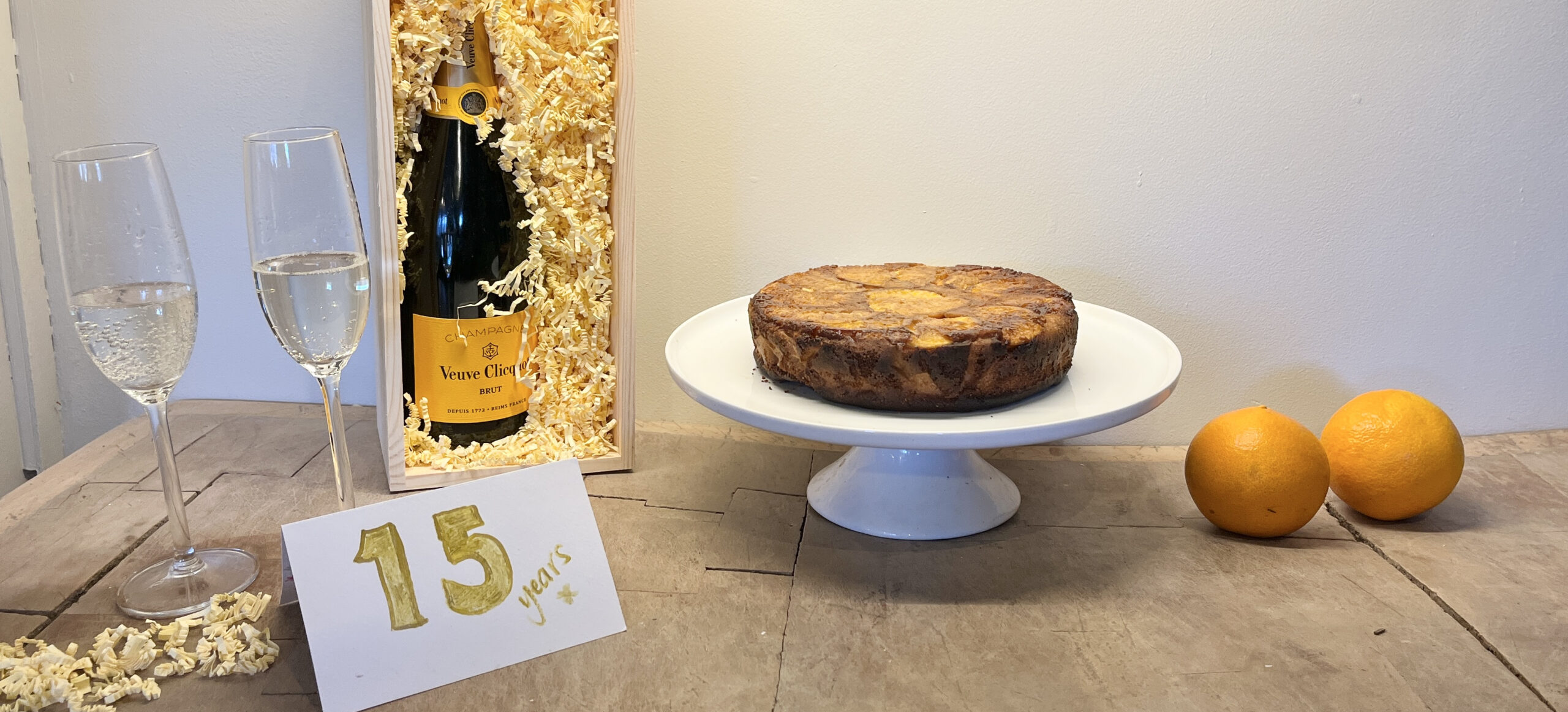 Marmalade House Design celebrates 15 years with a champagne bottle in a wooden box, a rustic cake on a pedestal, and oranges on a wooden table.