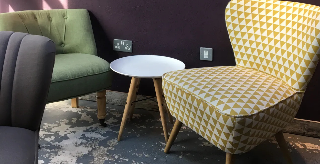 Sage green armchair and taupey sand with white geometric chairs around a round white side table against a deep purple wall, New Beginnings.